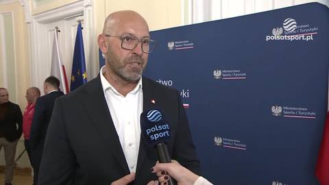 Jakub Rutnicki w okularach i garniturze udziela wywiadu przed niebieskim banerem z logo.