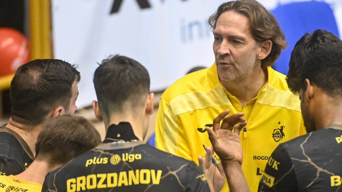 Trener w żółtej koszulce sportowej przemawia do grupy zawodników siatkówki w czarnych strojach, skupionych wokół niego.