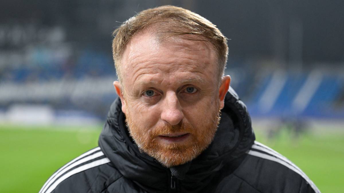 Portret mężczyzny w średnim wieku z rudą brodą, ubranego w czarną kurtkę z białymi paskami na ramionach.