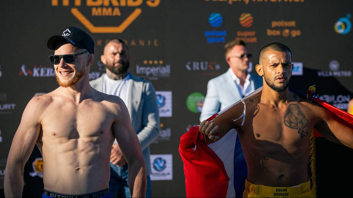 Dwóch mężczyzn stojących na scenie, prawdopodobnie przed walką MMA. Pierwszy od lewej ma na sobie czapkę i okulary przeciwsłoneczne, jest bez koszulki i ma widoczne mięśnie. Drugi od prawej również jest bez koszulki, ma tatuaż na klatce piersiowej i trzyma czerwoną szmatę. W tle widać logo Hybrid MMA i sponsorów.