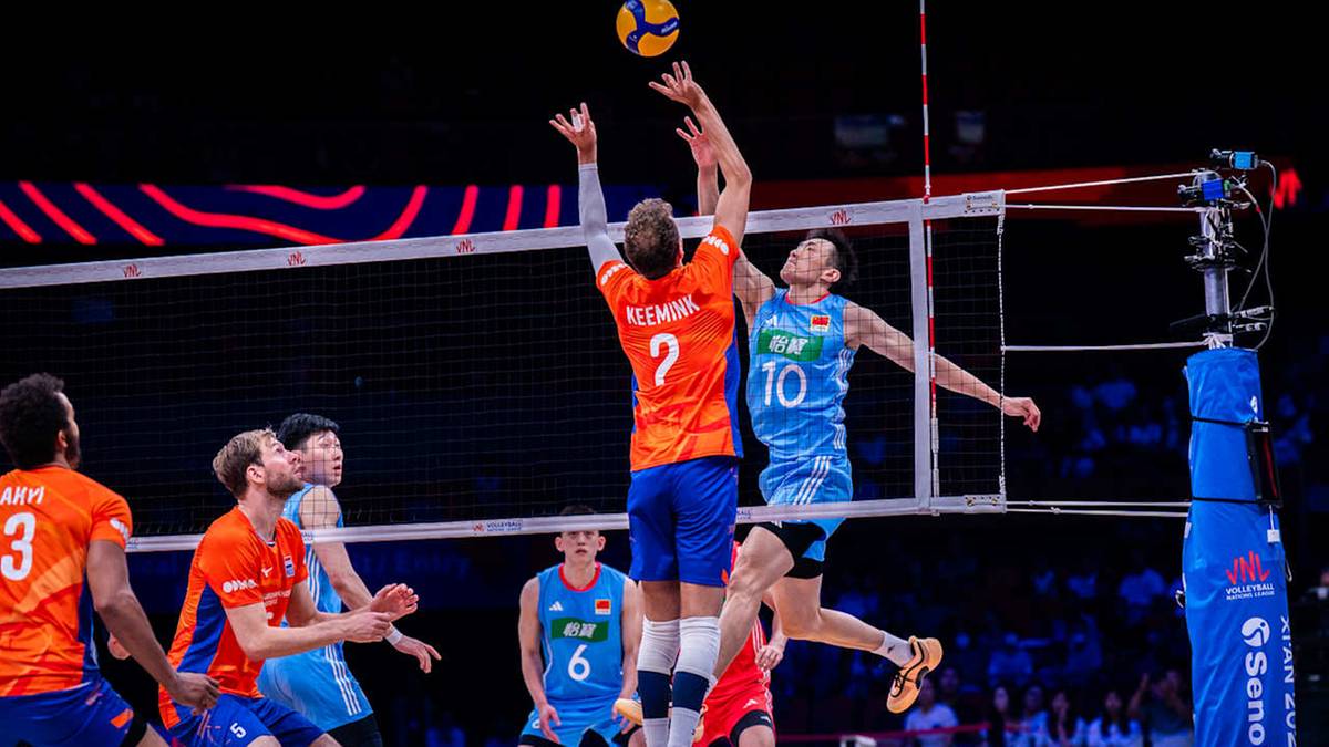 Gracz w niebieskim stroju skacze do ataku, próbując przebić piłkę nad siatką. Po drugiej stronie siatki dwaj gracze w pomarańczowych strojach blokują próbę ataku. Mecz siatkówki.