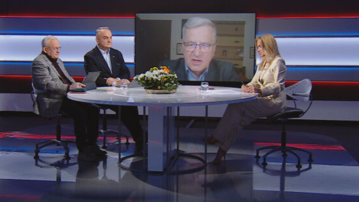 Trzy osoby siedzące przy okrągłym stole w studiu telewizyjnym, na ekranie widoczny czwarty mężczyzna.