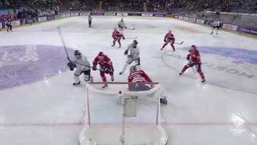 Zawodnicy drużyn IFK Helsinki i Sparta Praga grają w hokeja. Akcja toczy się pod bramką, gdzie jeden z zawodnik&oacute;w w czerwonym stroju strzela, a bramkarz w białym stroju pr&oacute;buje obronić. Inni zawodnicy w czerwonych i białych strojach znajdują się w pobliżu.