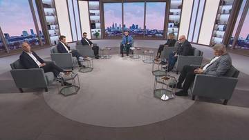 Grupa os&oacute;b siedzi w p&oacute;łokręgu na kanapach w studiu telewizyjnym z widokiem na panoramę miasta w tle.