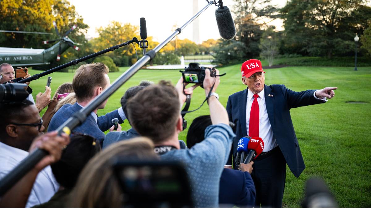 Donald Trump, ubrany w garnitur i czerwony kapelusz z napisem "USA", przemawia do grupy dziennikarzy z mikrofonami i aparatami fotograficznymi na tle Białego Domu.