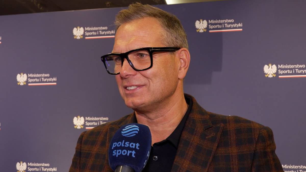 Mężczyzna w okularach i marynarce przed ścianką z logo Ministerstwa Sportu i Turystyki, trzymający mikrofon Polsat Sport.