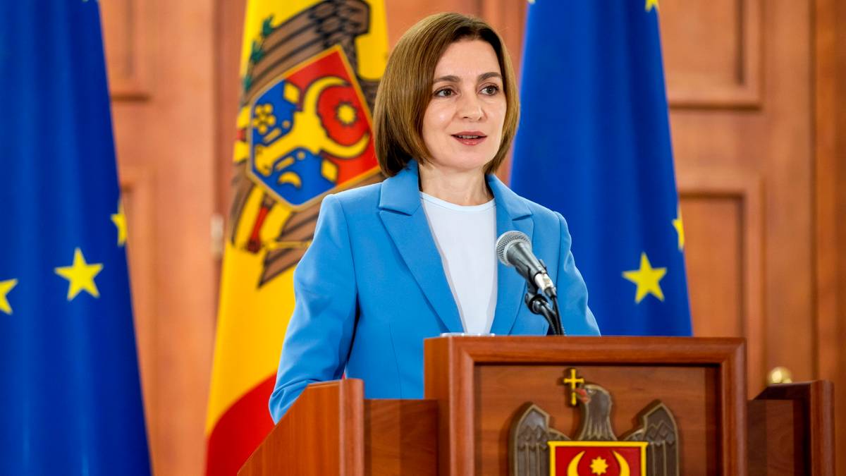 Kobieta w niebieskiej marynarce przemawia za mównicą z herbem, w tle powiewają flagi Mołdawii i Unii Europejskiej.