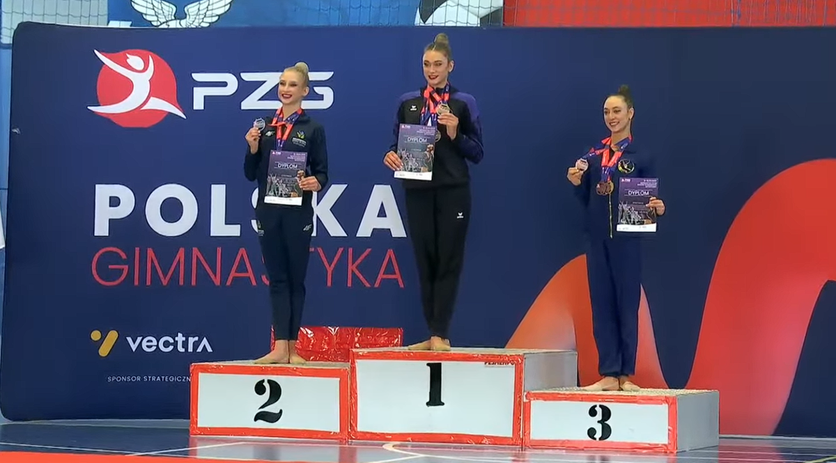 Trzy gimnastyczki stoją na podium po zawodach, z medalami na szyjach i dyplomami w rękach.