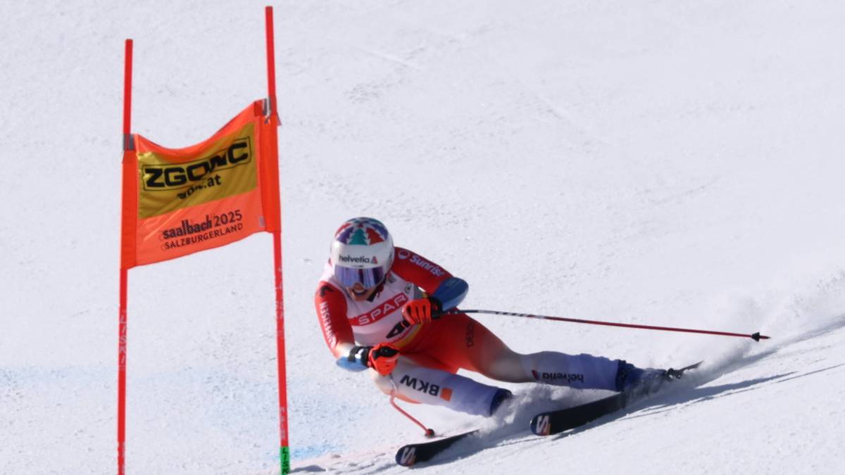 Narciarka alpejska w kombinezonie narciarskim i kasku jedzie na nartach po stoku, pochylona w zakręcie. Na tle pomarańczowa chorągiewka z logo.