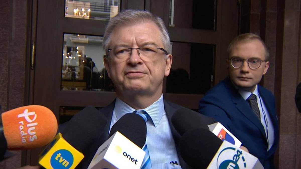 Mężczyzna w okularach i garniturze stoi przed mikrofonami.