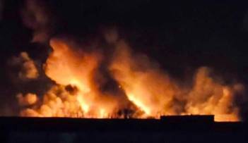 Widok nocny na płonącą rafinerię, z gęstymi kłębami dymu i intensywnymi płomieniami.