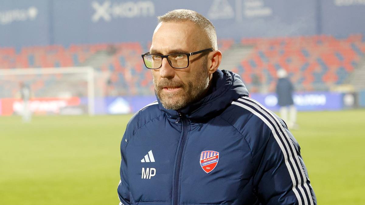Marek Papszun, trener Rakowa Częstochowa, w niebieskiej kurtce z logiem klubu, stoi na boisku piłkarskim.