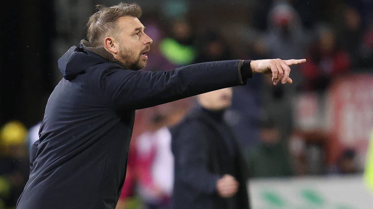 Trener piłkarski w ciemnej kurtce z kapturem wskazuje palcem w bok.