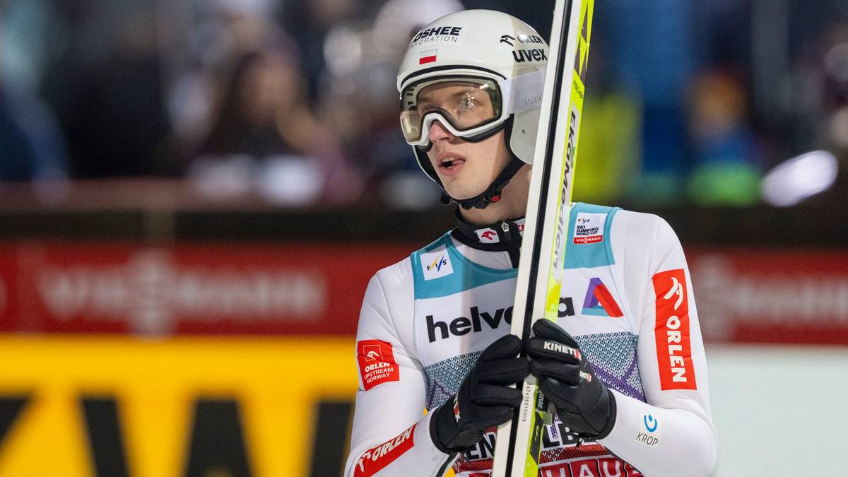 Skoczek narciarski w kasku i goglach, trzymający narty, w białym stroju z logo sponsor&oacute;w.