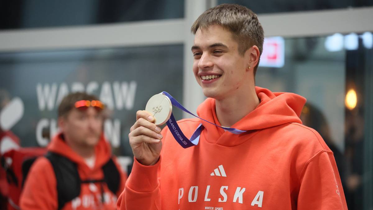 Uśmiechnięty młody mężczyzna w pomarańczowej bluzie z kapturem z napisem "POLSKA" trzyma medal na niebieskiej wstążce.