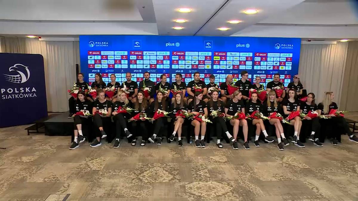 Drużyna siatkarek Polski siedzi w rzędzie na tle niebieskiej ściany z logo Polskiego Związku Piłki Siatkowej. Wszystkie zawodniczki mają na sobie czarne stroje sportowe z czerwonymi akcentami i uśmiechają się do kamery.