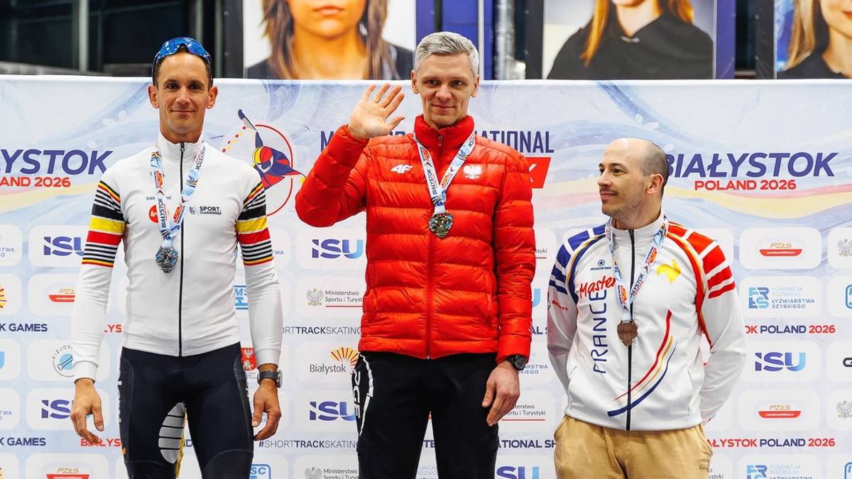 Trzej zawodnicy short tracku na podium, jeden z nich macha do publiczności.