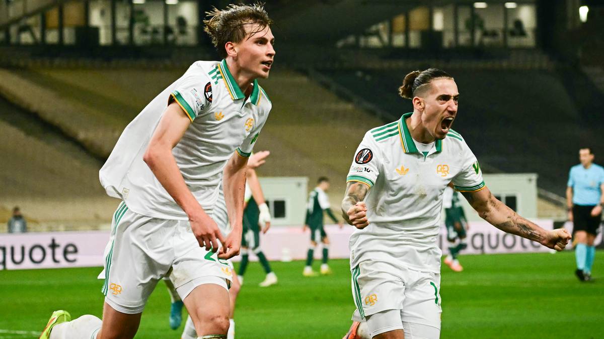 Dw&oacute;ch piłkarzy w białych strojach z zielonymi elementami cieszy się z bramki na boisku piłkarskim.