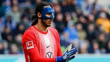 Bramkarz w czerwonej koszulce i niebieskiej masce na twarzy, trzymający rękawice.