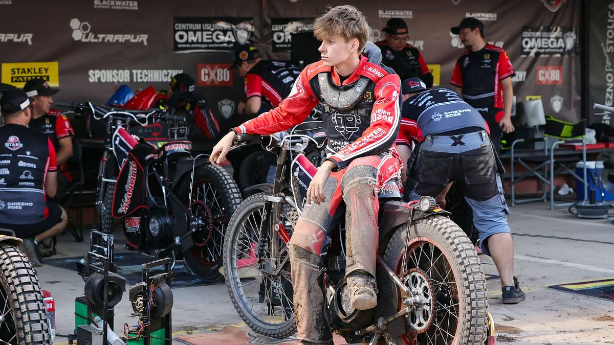 Młody żużlowiec w kombinezonie siedzi na motocyklu na torze żużlowym.