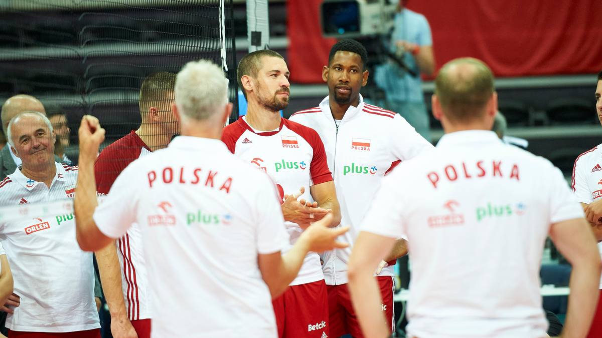 Grupa polskich siatkarzy w białych koszulkach z napisem "POLSKA" dyskutuje między sobą na hali sportowej.