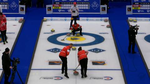 Gracze drużyny Polski i USA grają w curling. Dw&oacute;ch zawodnik&oacute;w z Polski stara się skierować kamień do domu, podczas gdy zawodnicy z USA obserwują grę. W tle widoczna jest tablica wynik&oacute;w i członkowie ekipy technicznej.