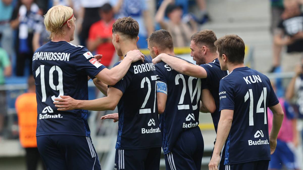 Grupa piłkarzy w granatowych strojach świętuje razem na boisku, niektórzy mają ręce na ramionach innych.