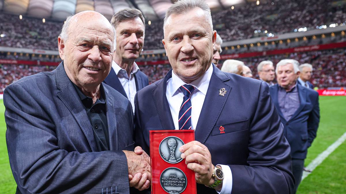 Dw&oacute;ch mężczyzn na boisku piłkarskim, jeden z nich ściska dłoń drugiemu i wręcza pamiątkową statuetkę.