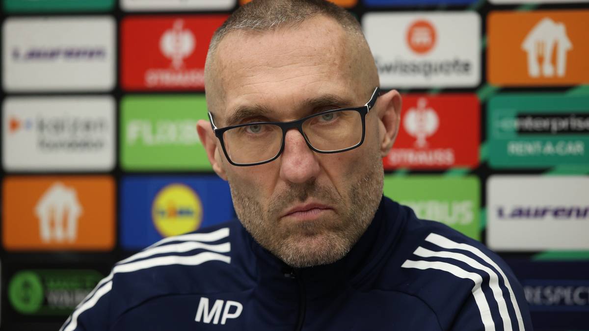 Portret mężczyzny w okularach, ubranego w sportową bluzę z widocznym herbem klubu i inicjałami "MP", na tle kolorowych paneli z logo sponsorów.