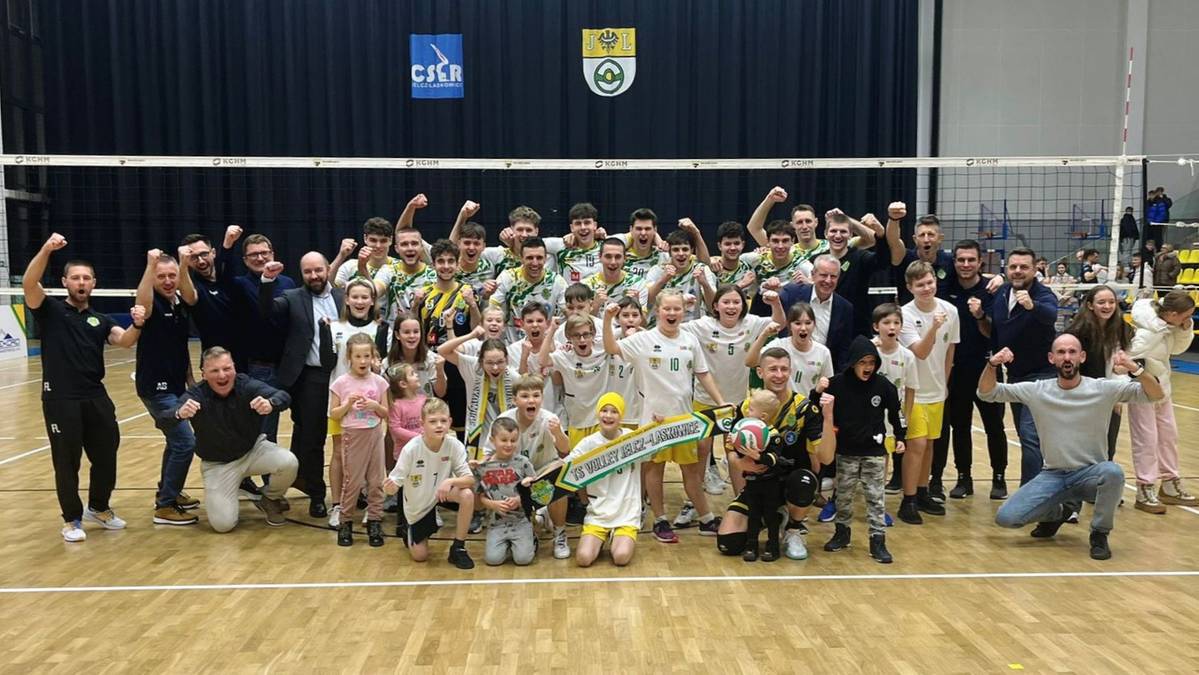 Drużyna siatkarska pozuje do radosnego zdjęcia grupowego na boisku z transparentem.