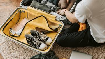 Osoba pakuje walizkę, obok leżą ubrania, buty i laptop.