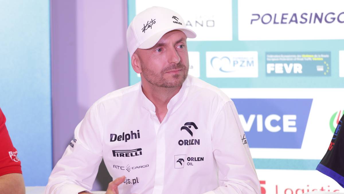 Kajetan Kajetanowicz, kierowca rajdowy, w białej koszuli z krótkim rękawem i białej czapce z daszkiem, siedzi podczas wydarzenia.