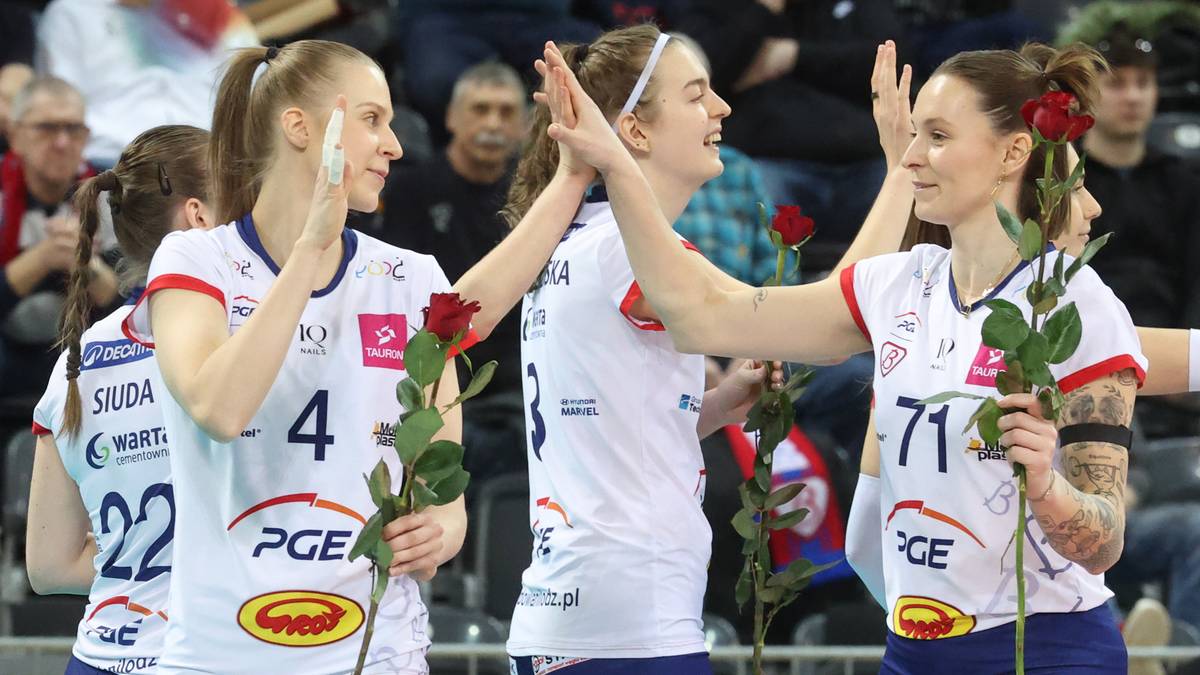 Trzy uśmiechnięte siatkarki w białych strojach sportowych z czerwonymi akcentami, trzymające czerwone r&oacute;że.