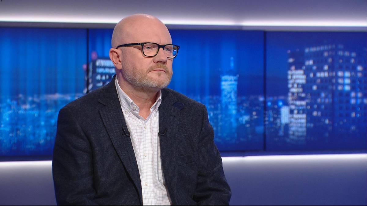 Maciej Duszczyk, wiceszef MSWiA, siedzi w studiu telewizyjnym przed ekranem z widokiem nocnego miasta.