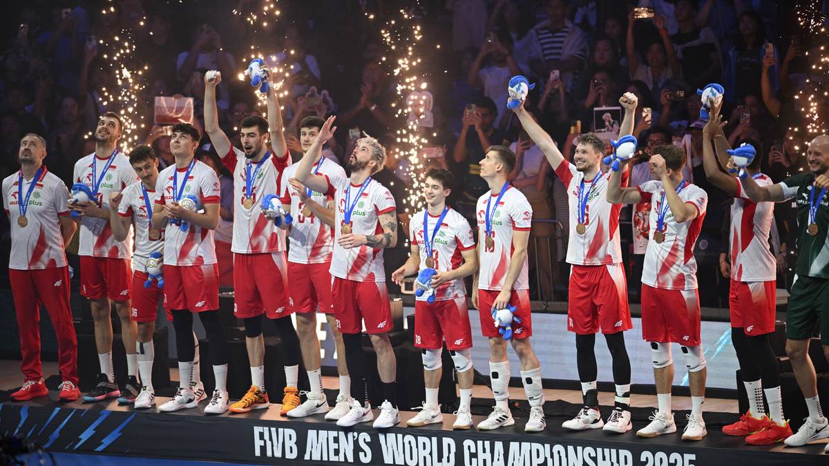 Drużyna siatkarzy w czerwonych strojach z medalami na szyjach, świętująca zwycięstwo na podium Mistrzostw Świata. W tle fajerwerki i tłum kibiców.