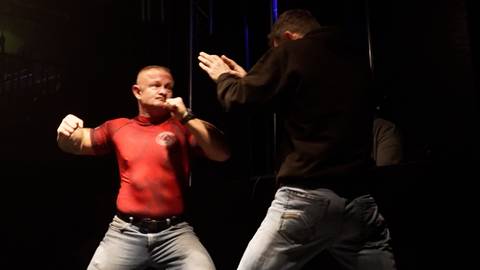 Dw&oacute;ch mężczyzn podczas ćwiczeń samoobrony w ciemnym pomieszczeniu. Jeden mężczyzna w czerwonej koszulce przyjmuje pozycję obronną, drugi w czarnej bluzie wykonuje ruch. Oświetlenie skupia się na ćwiczących.