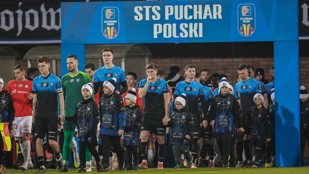 Grupa piłkarzy w niebieskich strojach wychodzi na boisko pod łukiem z napisem STS Puchar Polski.