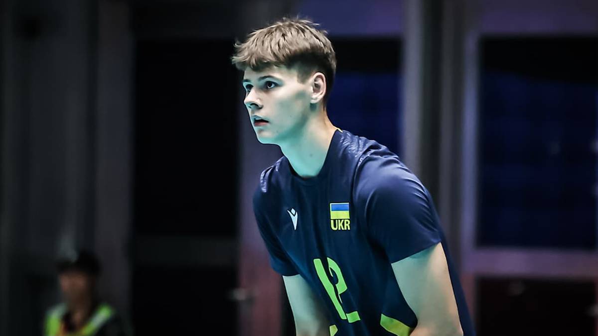 Młody mężczyzna w ciemnoniebieskiej koszulce z logo i napisem, z uwagą patrzy w bok.