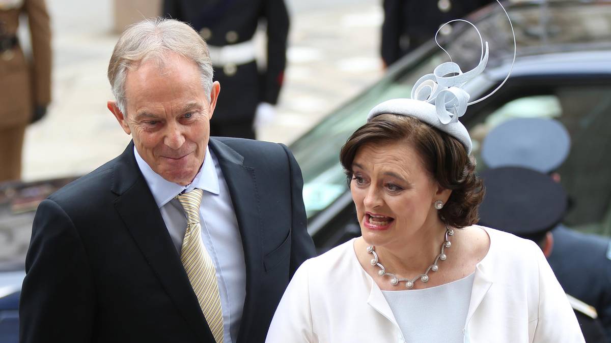 Cherie Blair w białej sukience i płaszczu oraz Tony Blair w garniturze idą obok siebie, wychodząc na zewnątrz.