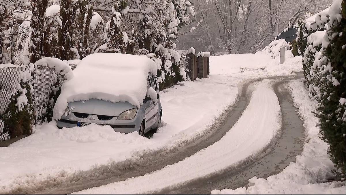 Samochód osobowy przykryty grubą warstwą śniegu, stojący na poboczu zaśnieżonej drogi. Po bokach widoczne są drzewa pokryte śniegiem.
