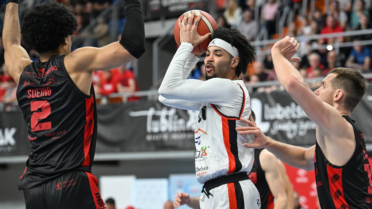 Trzech zawodników podczas meczu koszykówki. Jeden zawodnik z piłką w rękach, gotowy do rzutu. Dwóch zawodników drużyny przeciwnej próbuje go zablokować.