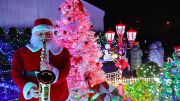 Postać w stroju Świętego Mikołaja gra na saksofonie przed choinką ozdobioną lampkami i innymi świątecznymi dekoracjami.