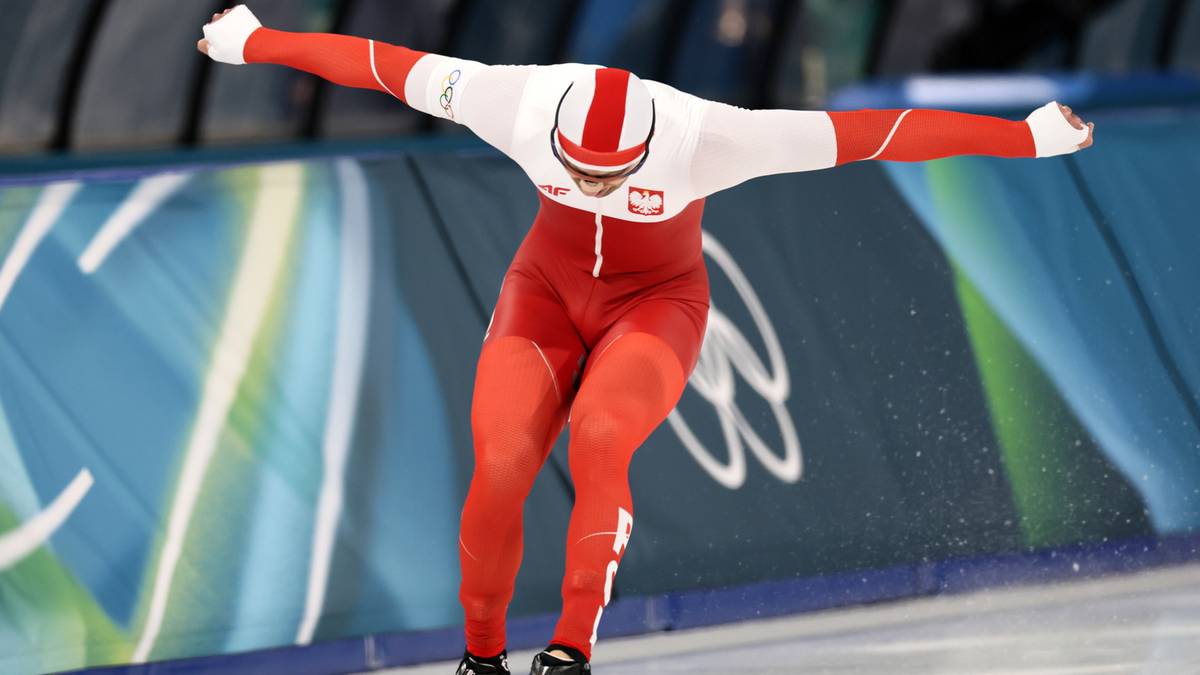 Polski panczenista w czerwono-białym stroju sportowym, szeroko rozłożonymi rękami i pochylonym ciałem, ślizga się po lodowisku podczas zawod&oacute;w.