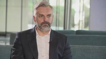 Mężczyzna z siwą brodą i wąsami, ubrany w marynarkę i koszulę, siedzi na kanapie. W tle nowoczesne wnętrze z przeszklonymi ścianami.