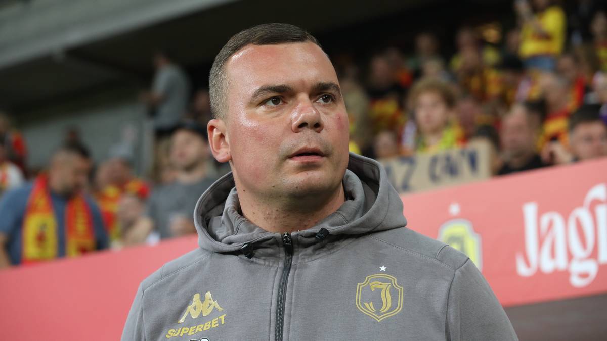 Zbliżenie na twarz trenera Jagiellonii Białystok w szarej bluzie z kapturem, z tyłu widownia stadionu.