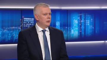 Mężczyzna w garniturze i krawacie m&oacute;wi, na tle widać ekran z nocnym widokiem miasta.