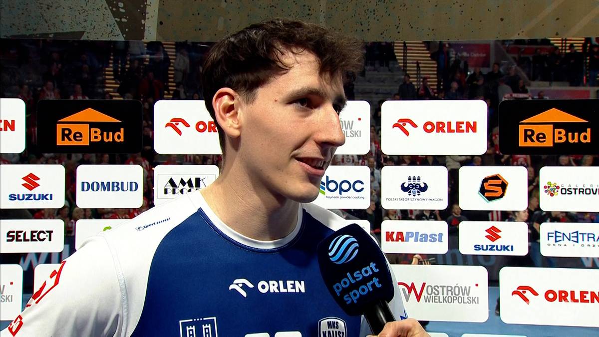 Młody mężczyzna w stroju sportowym, uśmiechnięty, trzymający mikrofon, na tle tablic z logotypami sponsorów.