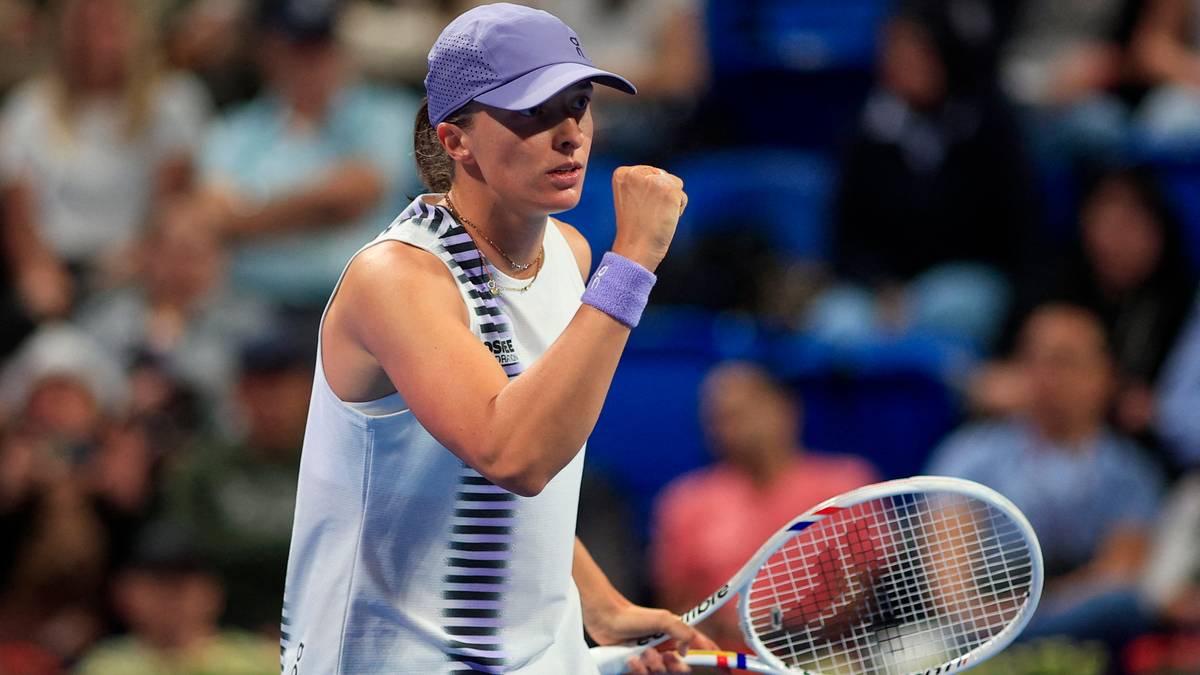 Iga Świątek w białej koszulce i fioletowej czapce, zaciskając pięść, cieszy się ze zwycięstwa w meczu tenisowym.