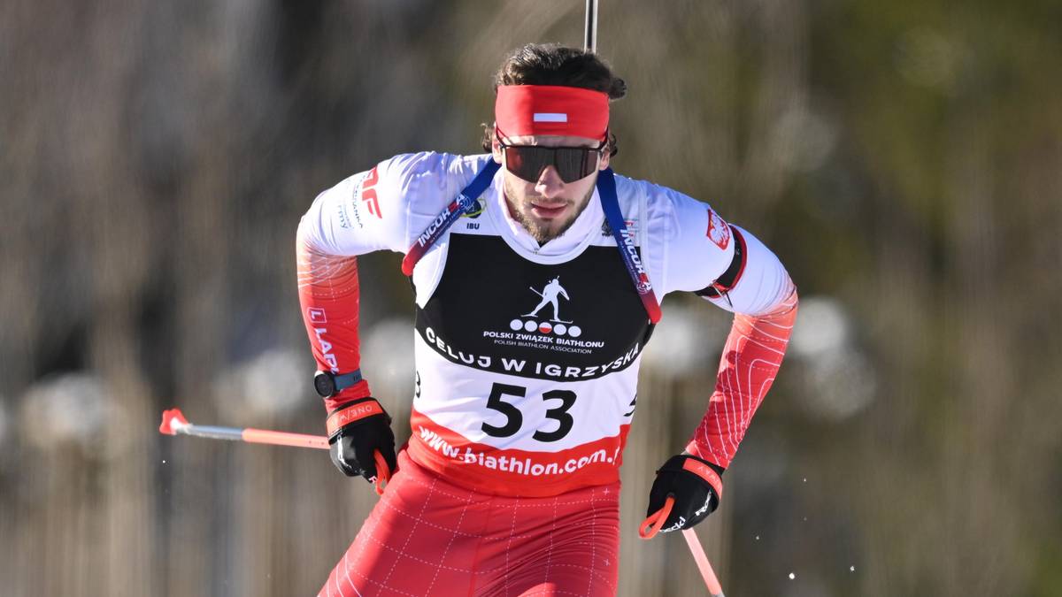 Biathlonista w czerwonym stroju narciarskim z karabinem na plecach stoi na ośnieżonej trasie.