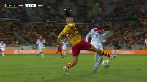 Piłkarz Jagiellonii Białystok w ż&oacute;łtej koszulce i czerwonych spodenkach pr&oacute;buje kopnąć piłkę, podczas gdy przeciwnik w białej koszulce stara się mu przeszkodzić.